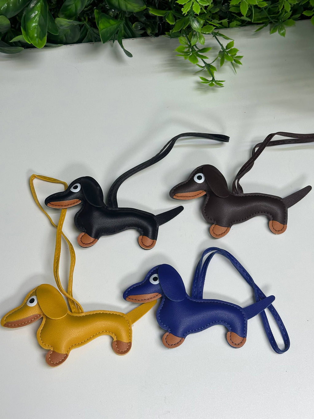 Three leather dachshund keychains in black, blue, and yellow on a white surface with greenery in the background.