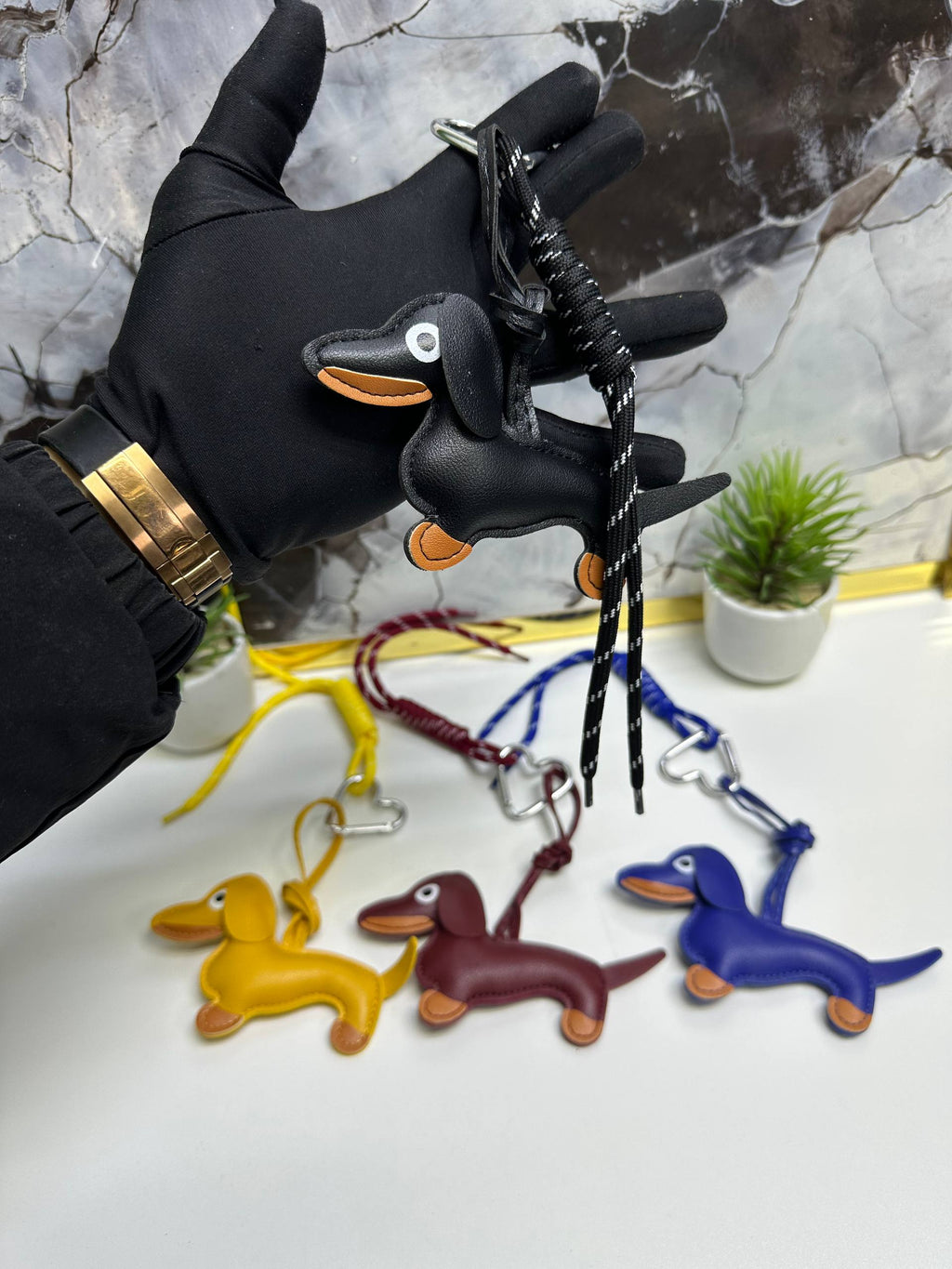 Black glove holding a black dachshund keychain with other colored dachshund keychains on a white surface.